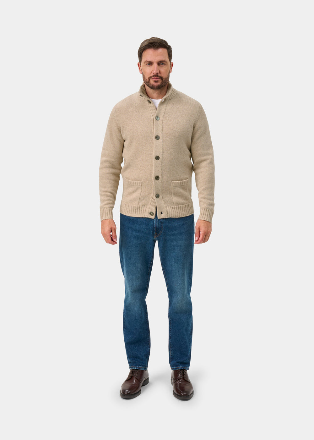 A man wears the Landford Lambswool Buttoned Mock Neck Jumper in Linen with blue jeans and brown shoes on white.