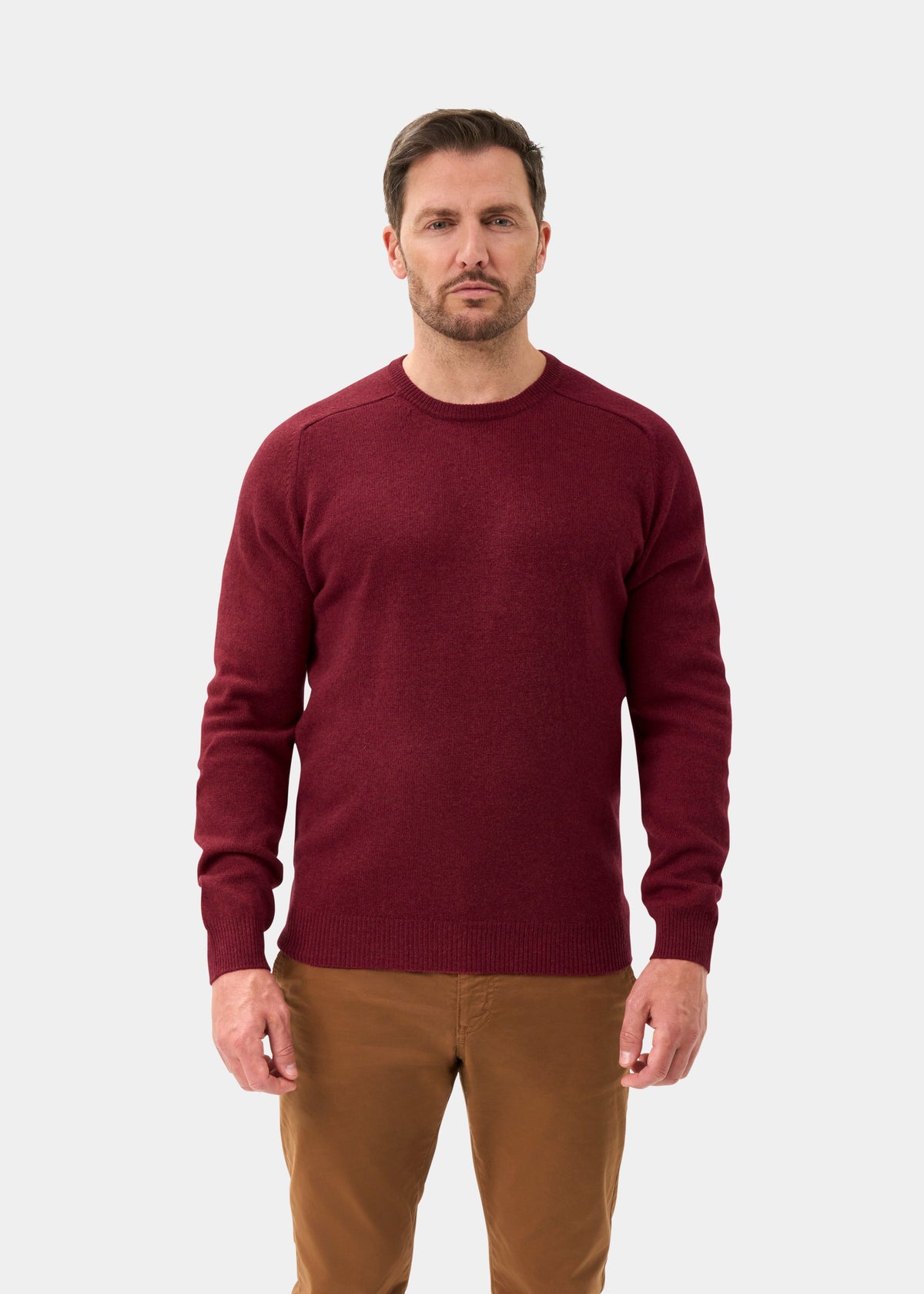 Man in Lenzie Lambswool Crew Neck Jumper in Garnet, regular fit, and brown pants stands against a light background.