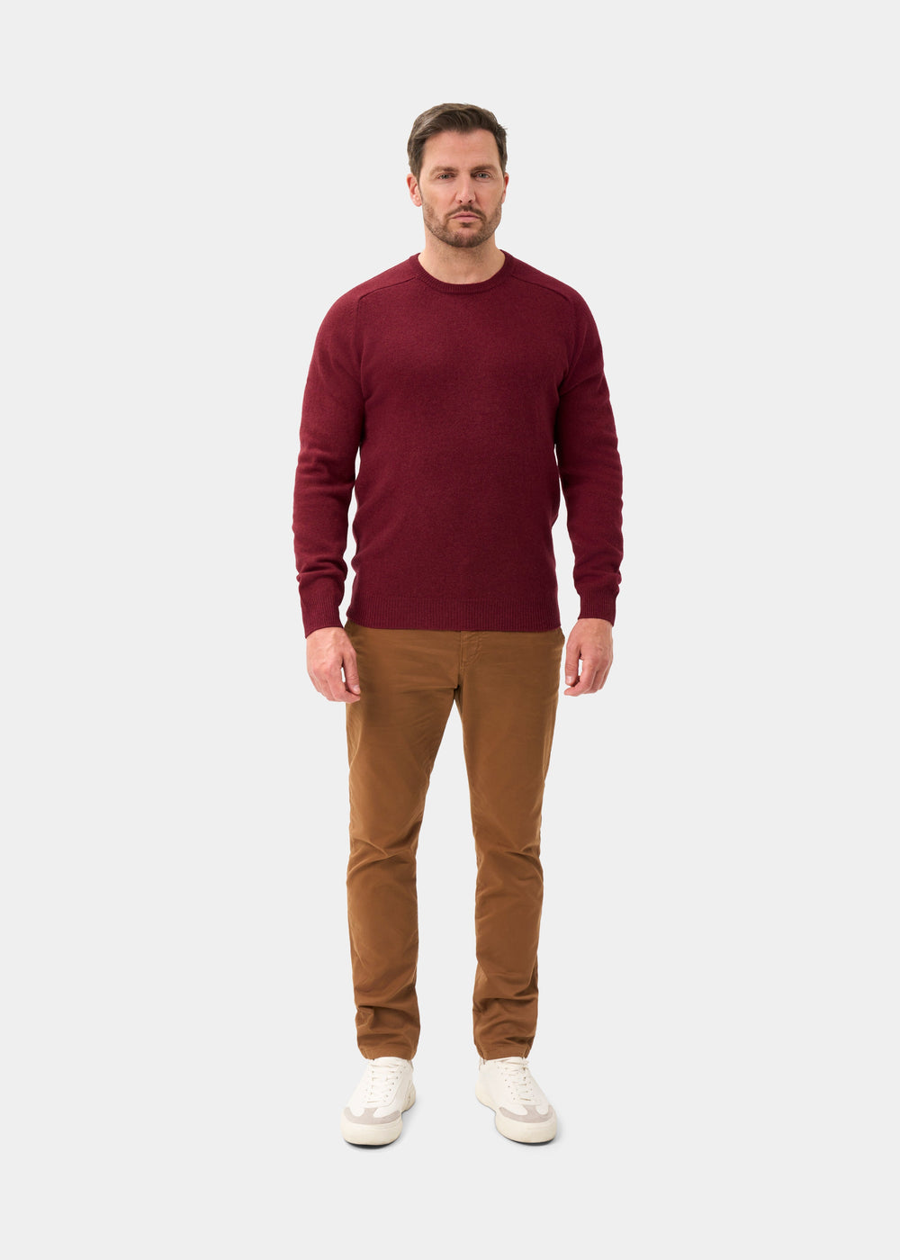 Man in Lenzie Lambswool Crew Neck Jumper in garnet, brown pants, and white sneakers against a plain background.
