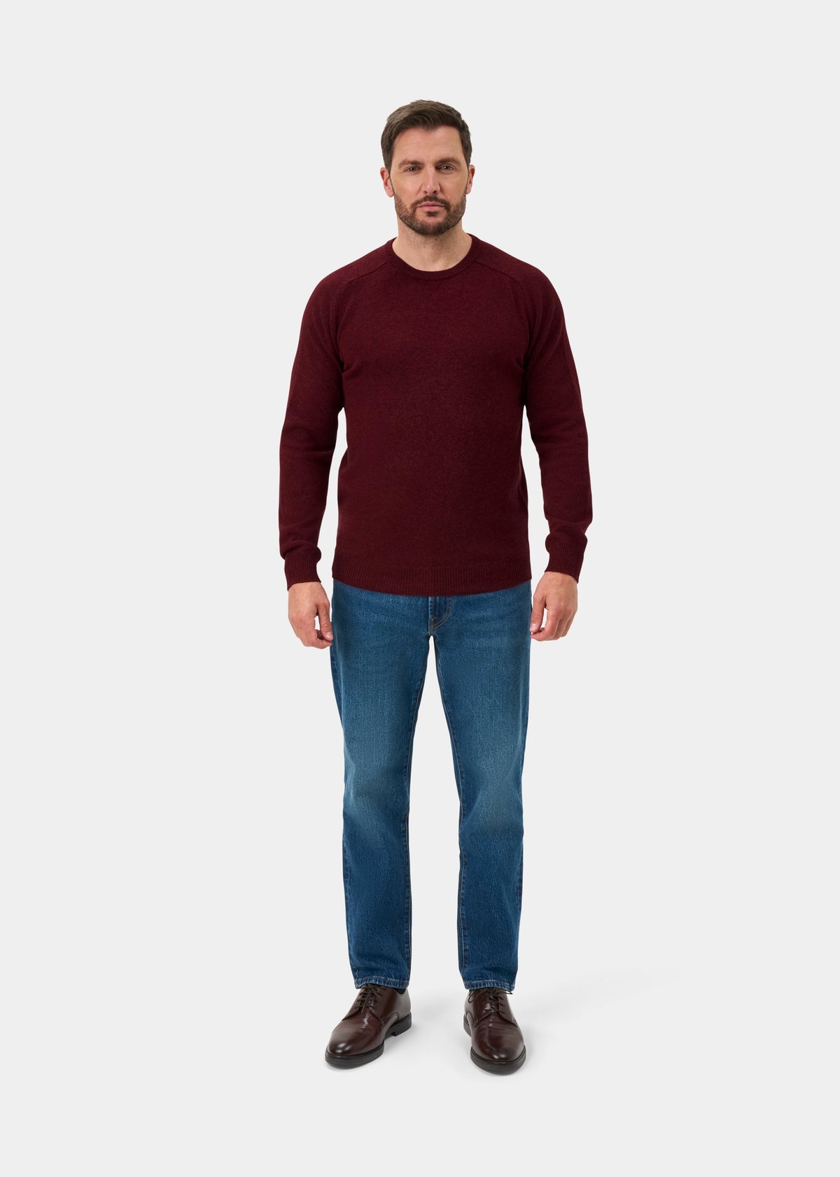Man in a red lambswool crew neck jumper, blue jeans, and brown shoes stands facing forward against a plain backdrop.