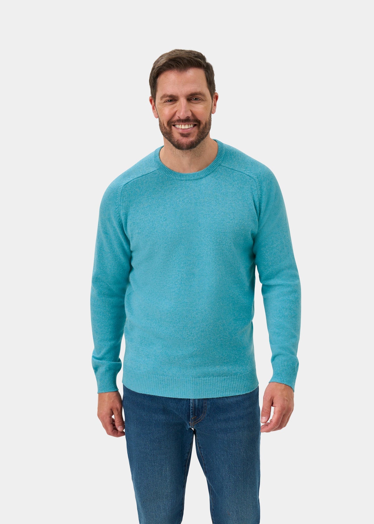 Man smiles in Lenzie Lambswool Crew Neck Jumper in Topaz and blue jeans, against a plain white background.