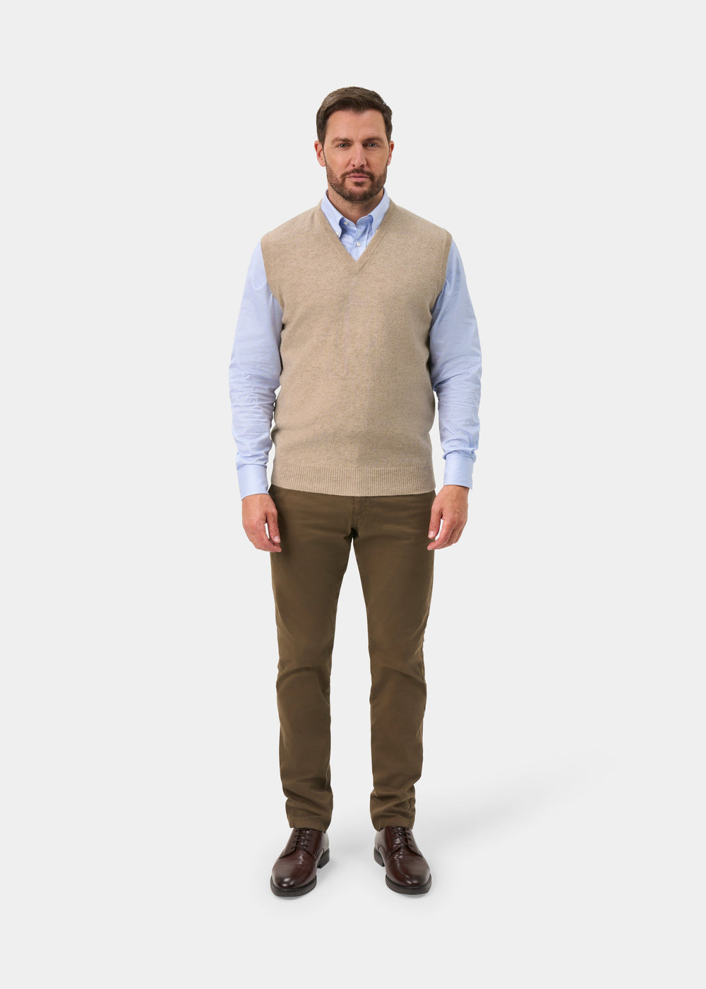 A man modeling a beige v-neck knit sweater vest over a light blue dress shirt, paired with olive brown trousers and dark brown leather shoes, against a light gray studio background.