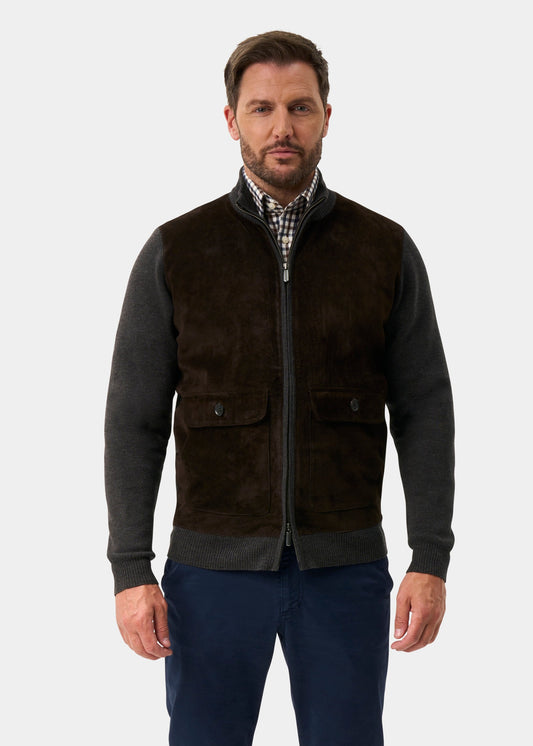 A man modeling a dark brown suede vest over a dark gray zip-up sweater, paired with navy blue trousers and brown leather shoes, against a light gray studio background.